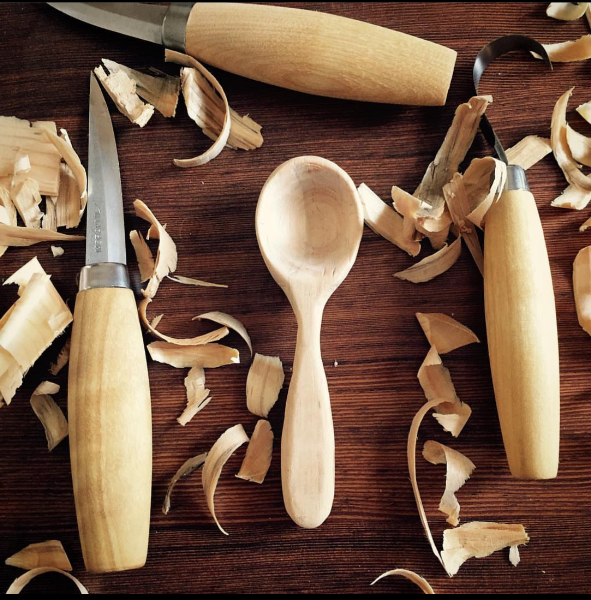 A carved spoon witht he carving knives around it and also wood shavings