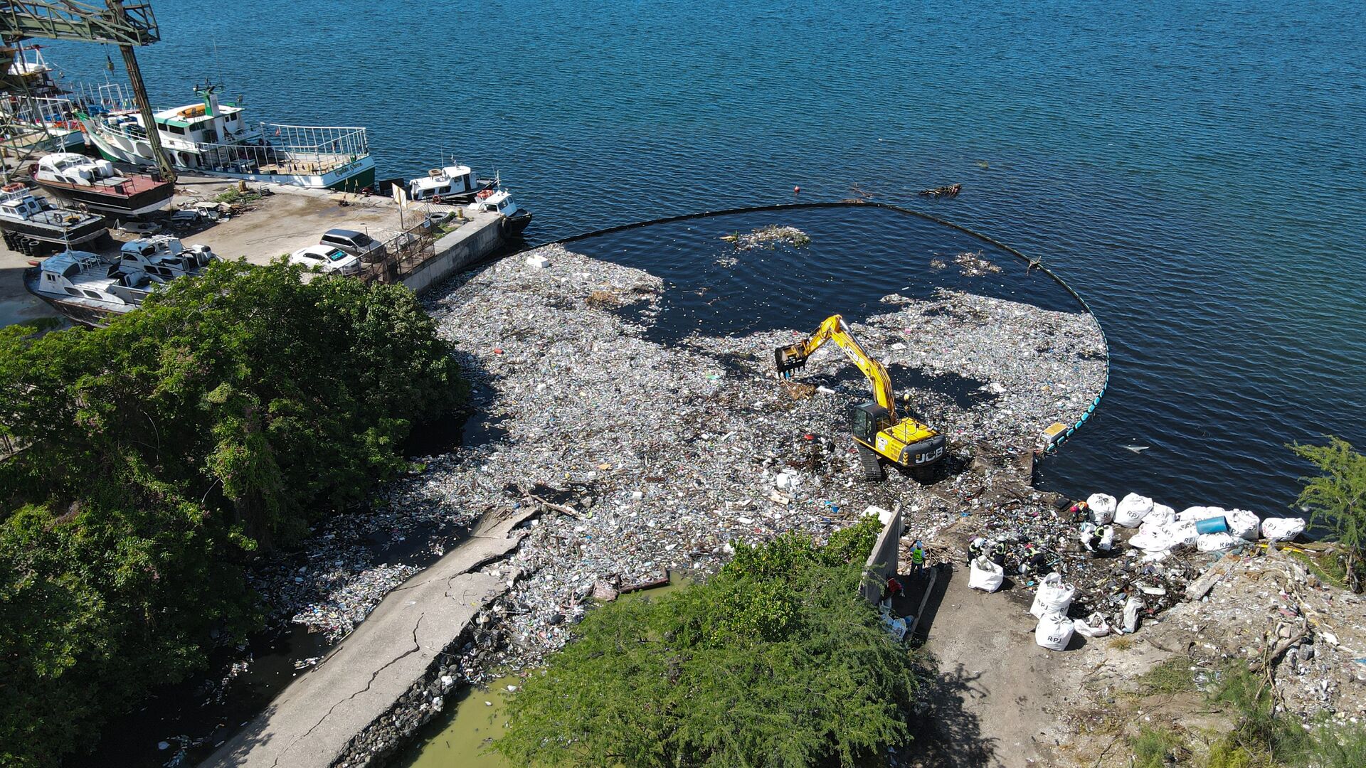 An excavator removing trapped waste from the Kingston Harbour shoreline, with a floating barrier holding back debris offshore.