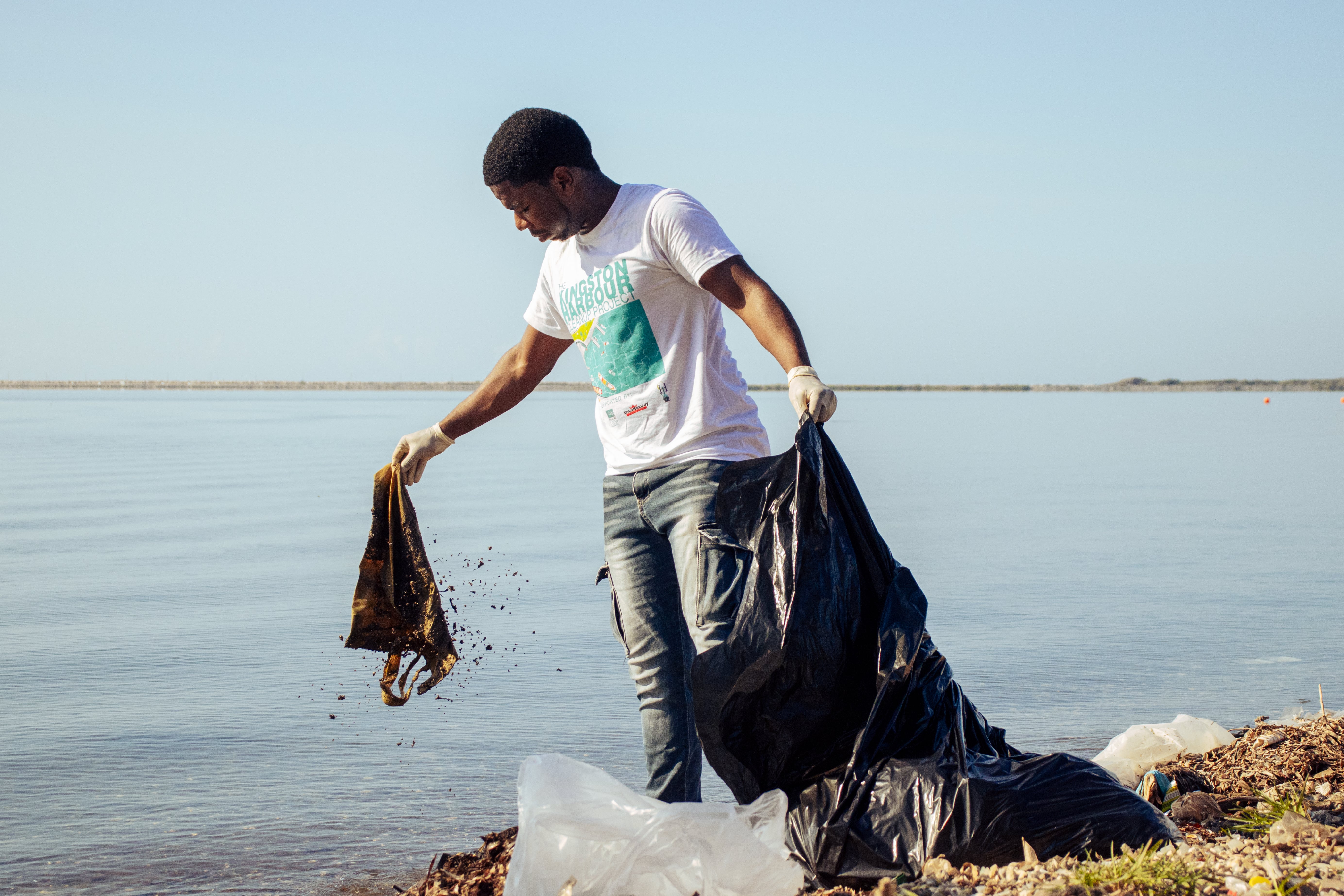 A volunteer wearing gloves lifting waste from the shoreline into a black bag during a beach cleanup.