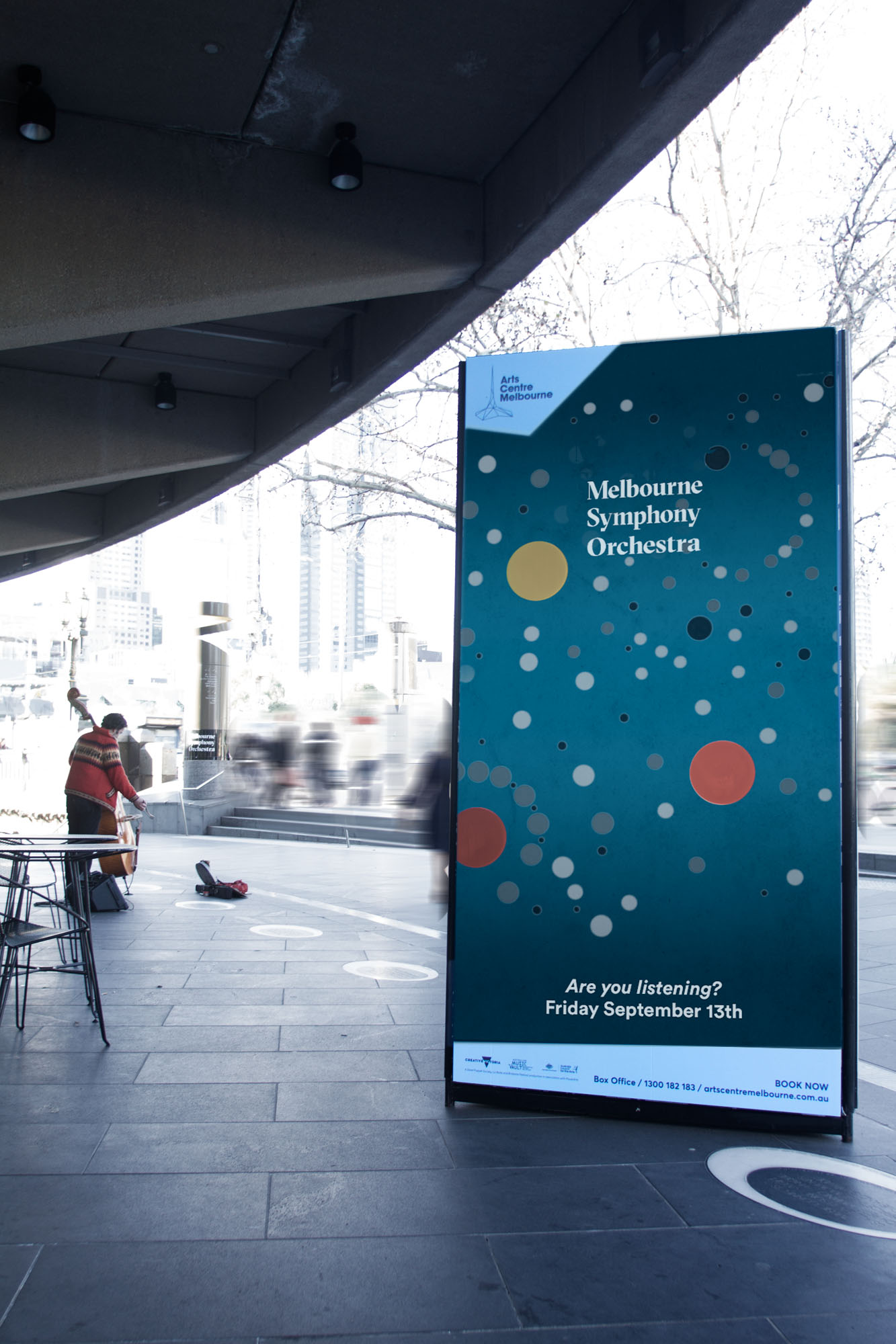 A digital poster board with a sound visualisation and a man playing the Cello in the background outside Hamer Hall, Melbourne
