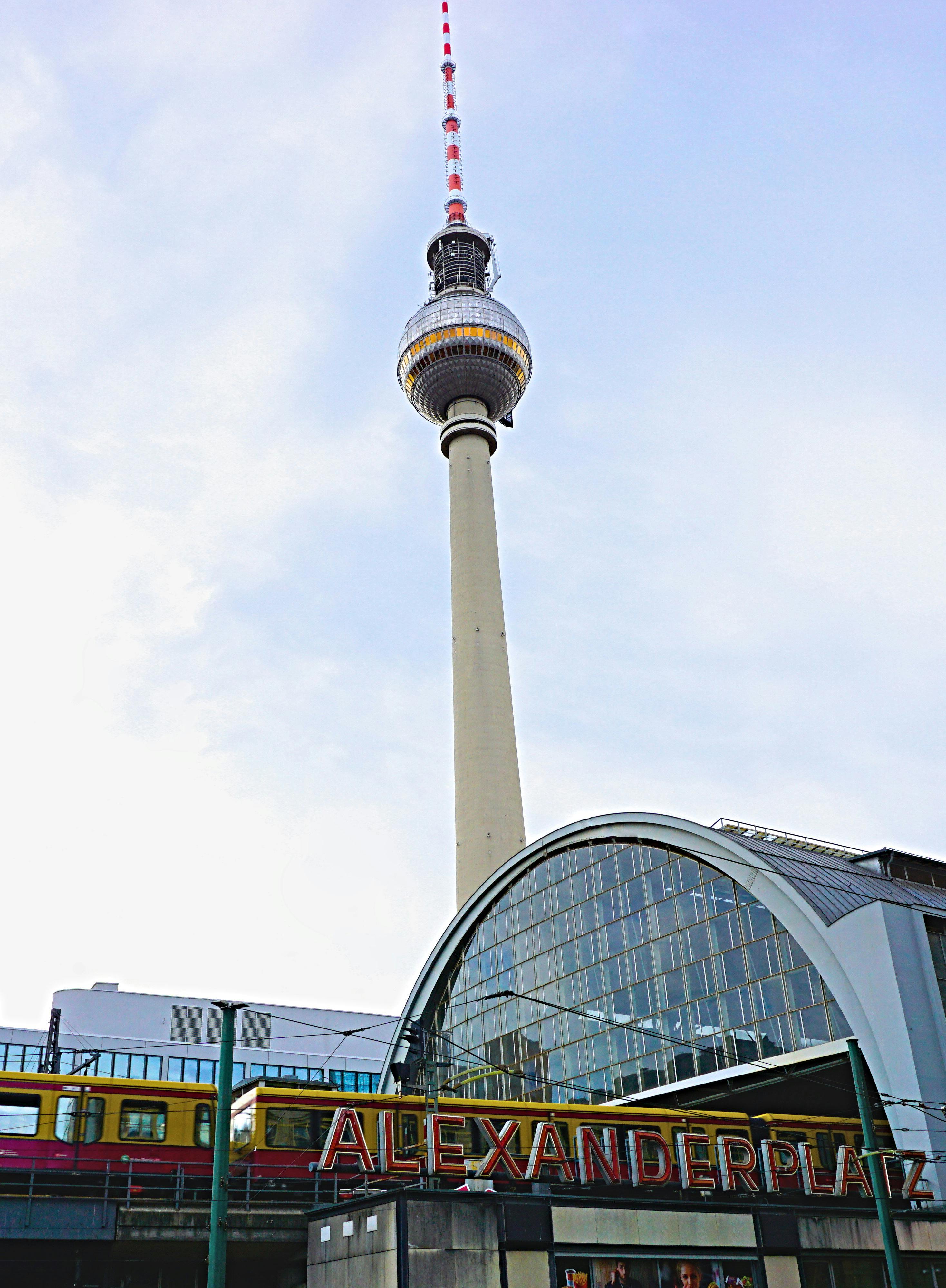 Das Bild zeigt den Berliner Fernsehturm, aufgenommen vom Alexanderplatz aus. Im Vordergrund ist das markante, gebogene Glasdach des S-Bahnhofs Alexanderplatz zu sehen, darüber verläuft eine gelb-rote S-Bahn. Der Fernsehturm ragt zentral in den Himmel, dessen helle, leicht bewölkte Färbung eine freundliche und klare Stimmung erzeugt. Das ikonische „ALEXANDERPLATZ“-Schild ist gut lesbar und vermittelt einen typischen Berliner Stadteindruck.