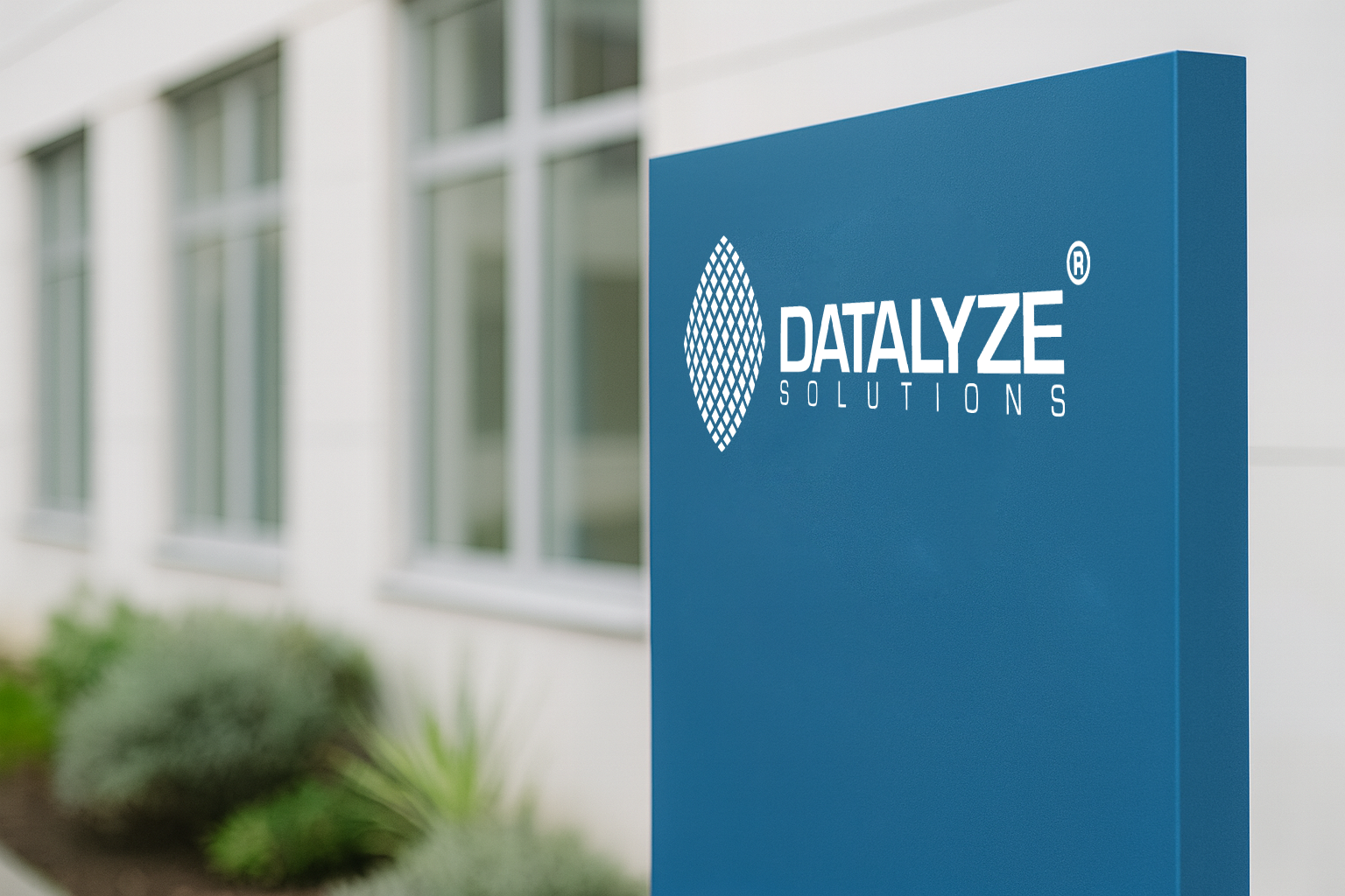 Das Bild zeigt ein fotorealistisches, modernes Firmenschild vor einem Bürogebäude. Das Schild ist rechteckig, in einem satten Blau gehalten, und trägt mittig das Original-Datalyze-Solutions-Logo in Weiß – mit dem charakteristischen Rautenmuster-Symbol links und der klaren Schrift „DATALYZE SOLUTIONS“ rechts daneben.