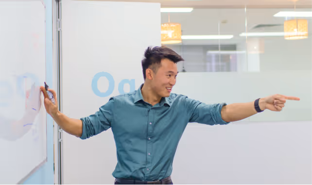 Young Cognito tutor in teal shirt smiling while standing by a whiteboard in a classroom.