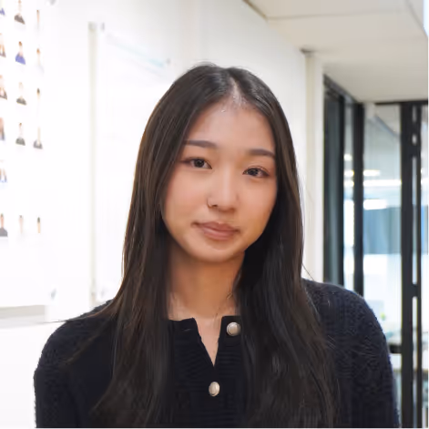 Young woman with long dark hair wearing a black cardigan standing indoors in a modern office space.
