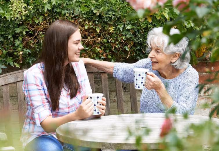 junge Frau lacht zusammen mit einer älteren Frau. Beide haben Tassen in der Hand und trinken Tee