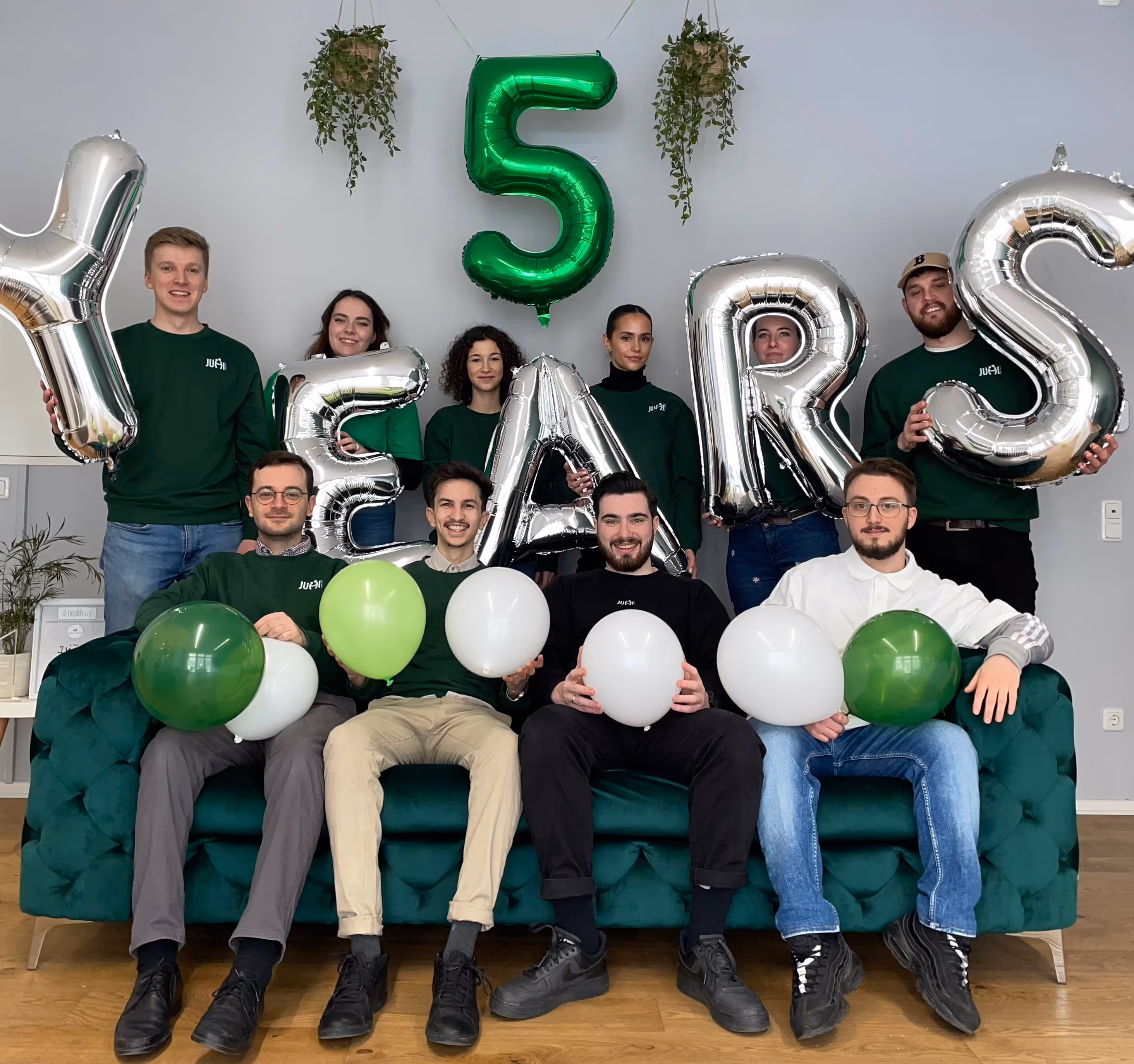 Group of ten young adults sitting and standing behind a green couch holding silver balloons spelling 'YEARS' and a green balloon shaped as number 5.