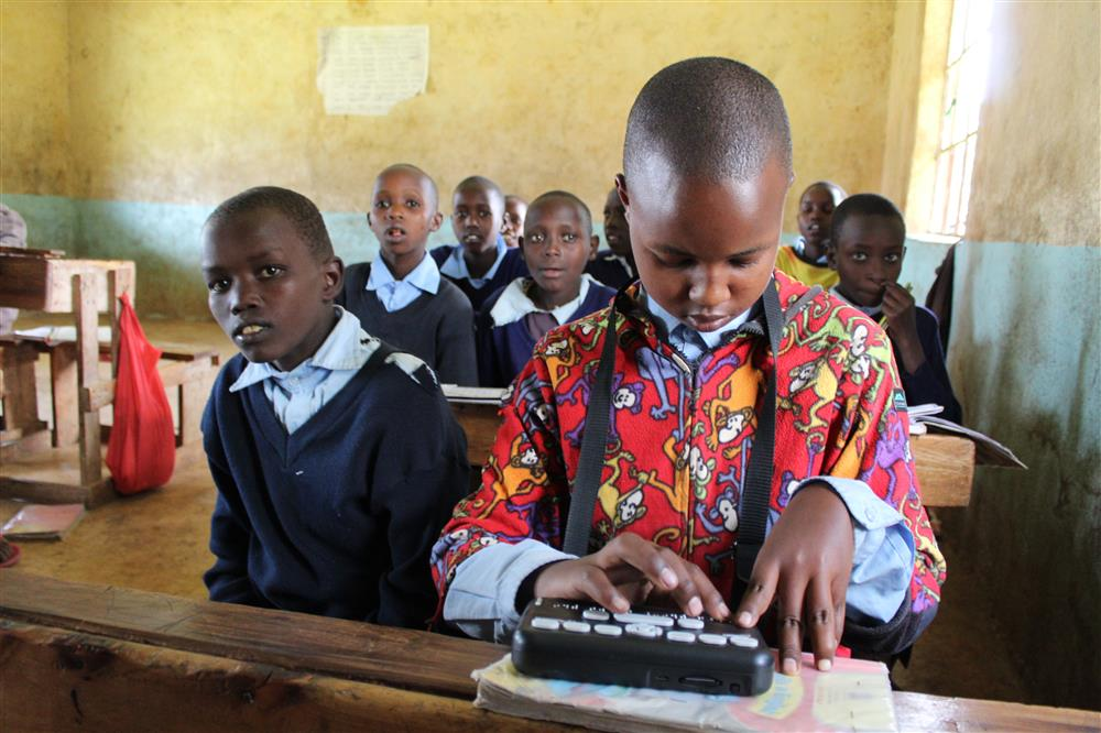 A visually impaired student in a colorful shirt uses a braille device at a classroom desk, while other students in uniform sit attentively behind.
