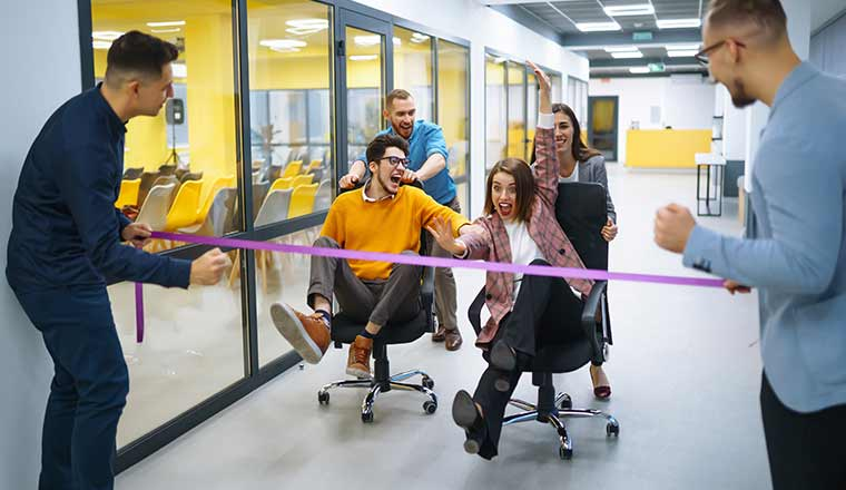 Office employees participating in a fun team-building race, pushing each other on chairs through a hallway and breaking a finish-line ribbon together.
