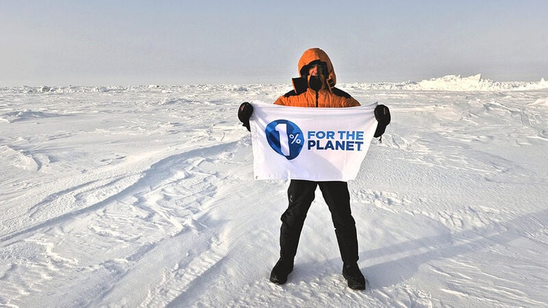 Person wearing an orange jacket standing on a snowy landscape holding a banner that reads “1% for the Planet.”