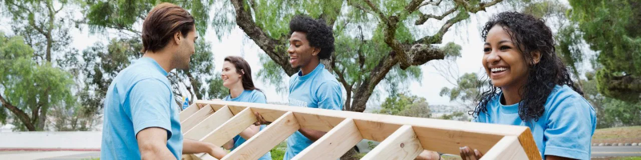 Group of corporate volunteers in blue shirts smiling and working together outdoors to carry wooden planks as part of a community service project.