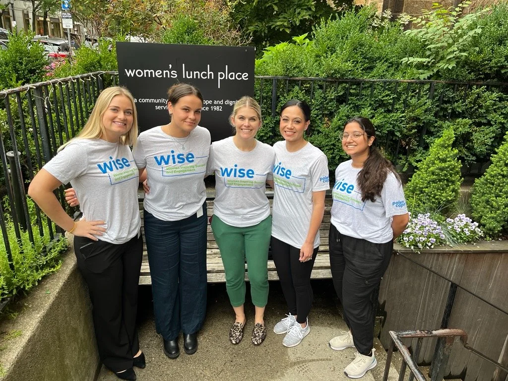 Group of women wearing WISE T-shirts standing outdoors near a Women’s Lunch Place sign, smiling together in a community setting.