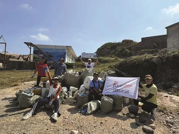 Volunteers pose outdoors with collected trash bags after a coastal cleanup, holding a Picking China banner under a clear sky.