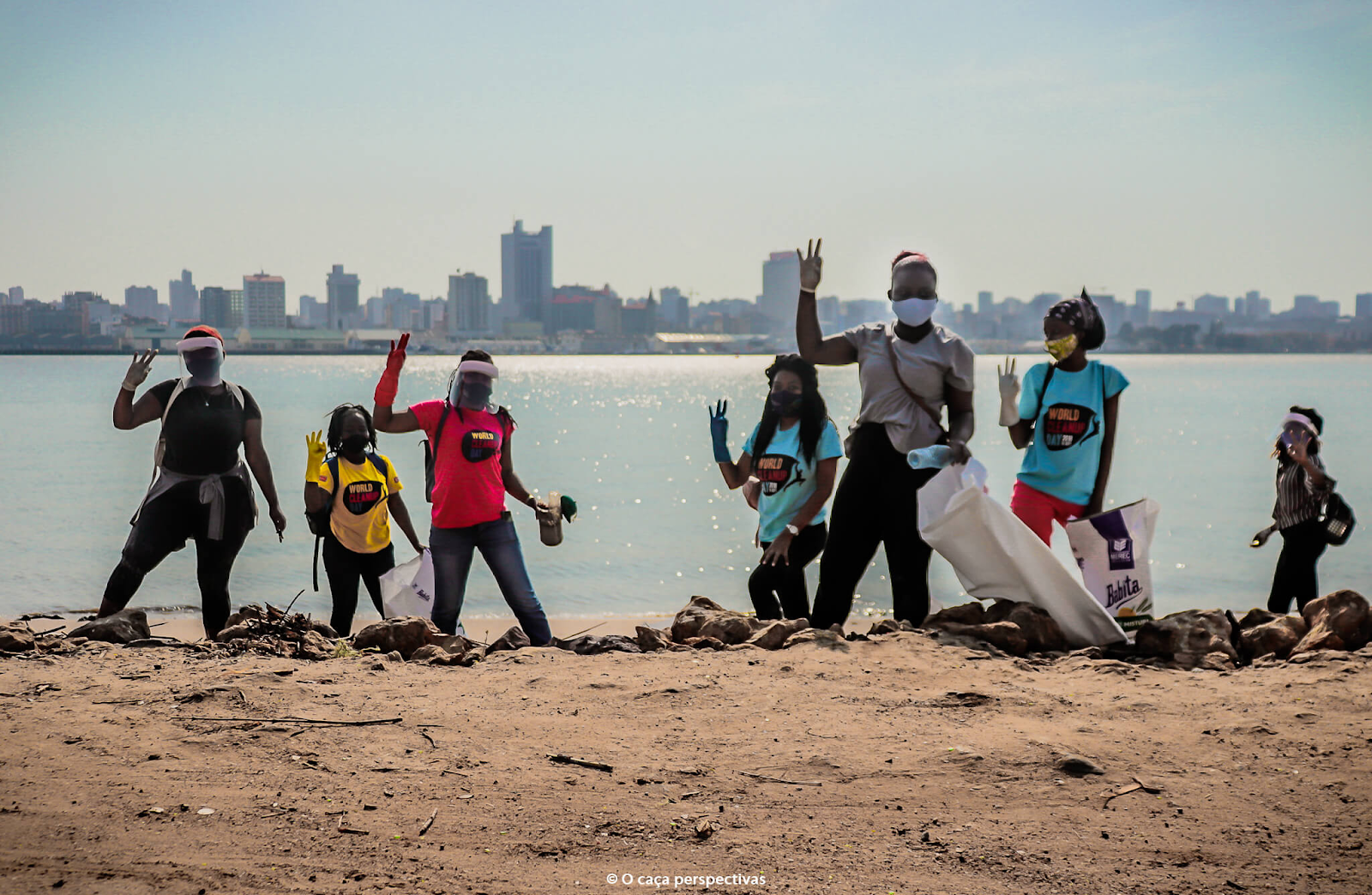 Volunteers wearing masks and gloves stand along a shoreline holding trash bags, waving after a beach cleanup with a city skyline in the background.