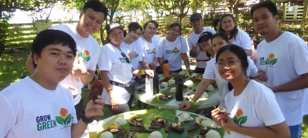A group of volunteers in Grow Green T-shirts gather outdoors around a table, sharing food and smiling during a community activity.