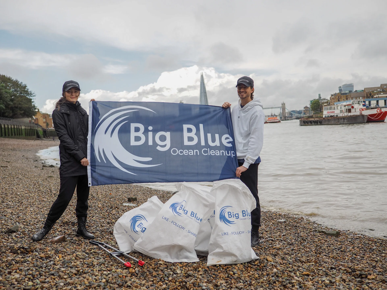 Two volunteers stand on a riverside holding a Big Blue Ocean Cleanup banner, with collected waste bags and a city skyline behind them.