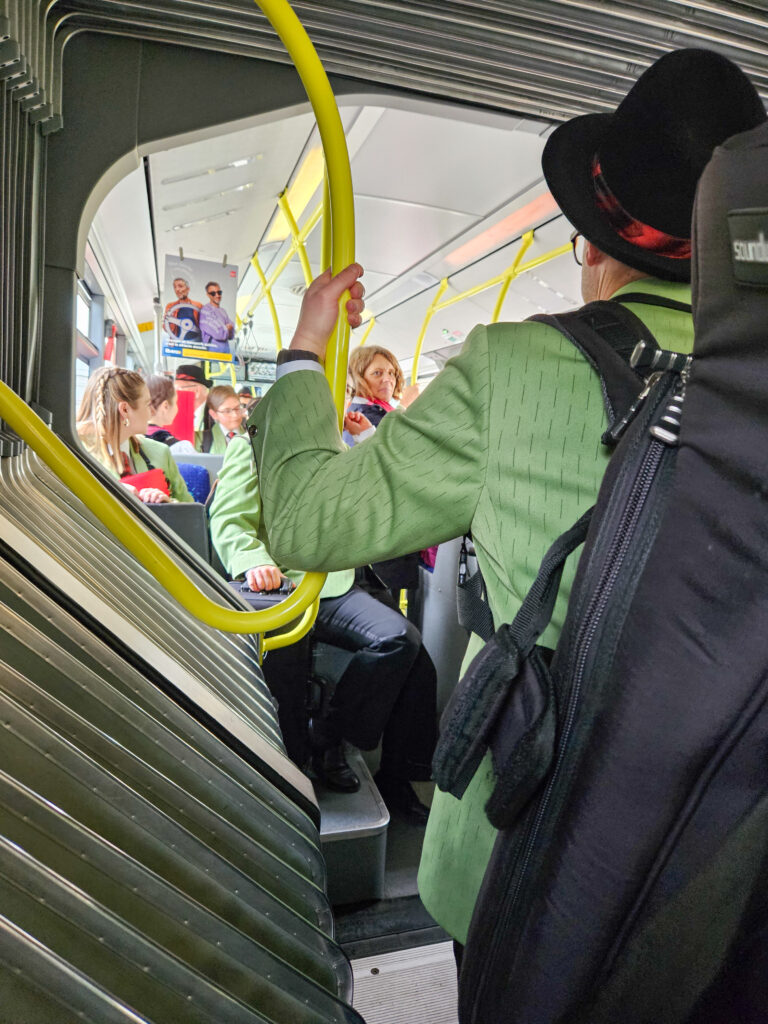 Musikant mit Instrumentenkoffer am Rücken von hinten sichtbar, vorne im Bus hat es noch mehr Musikanten in lindengrünen Uniformen