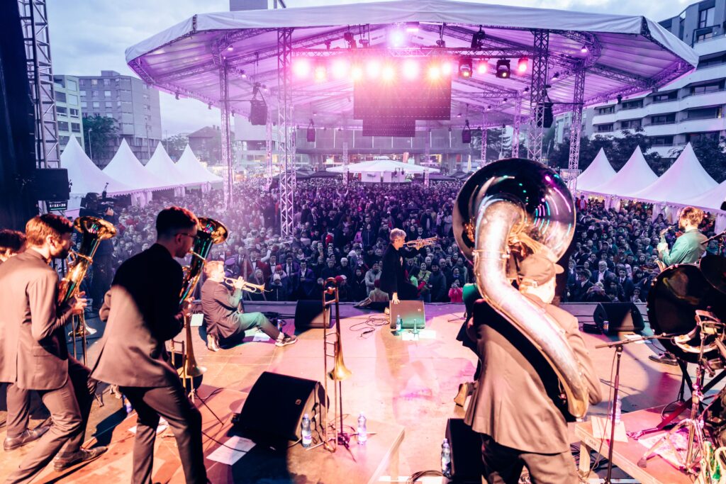 Blick von der Bühne mit Band, im Vordergrund ein Sousaphonspieler von hinten, auf die Massen unter dem Paragu