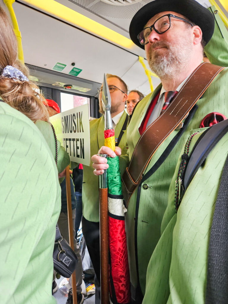 im Shuttlebus unterwegs mit dem Fähnrich - er trägt die Fahne - bei sich und Mitgliedern der Vereinsmusik Kriegstetten in lindengrüner Uniform und mit Täfeli für die Marschmusik