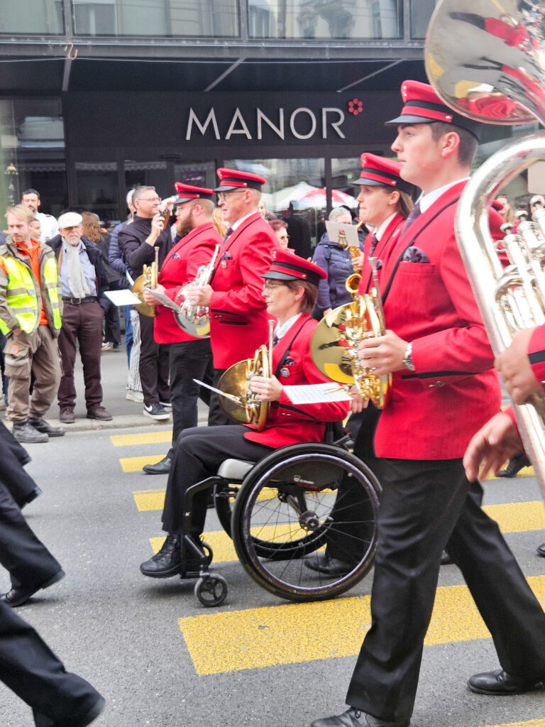 Musikanten in roten Uniformen mit Instrumenten marschierend, eine Hornistin wird im Rollstuhl sitzend von einer Frau geschoben