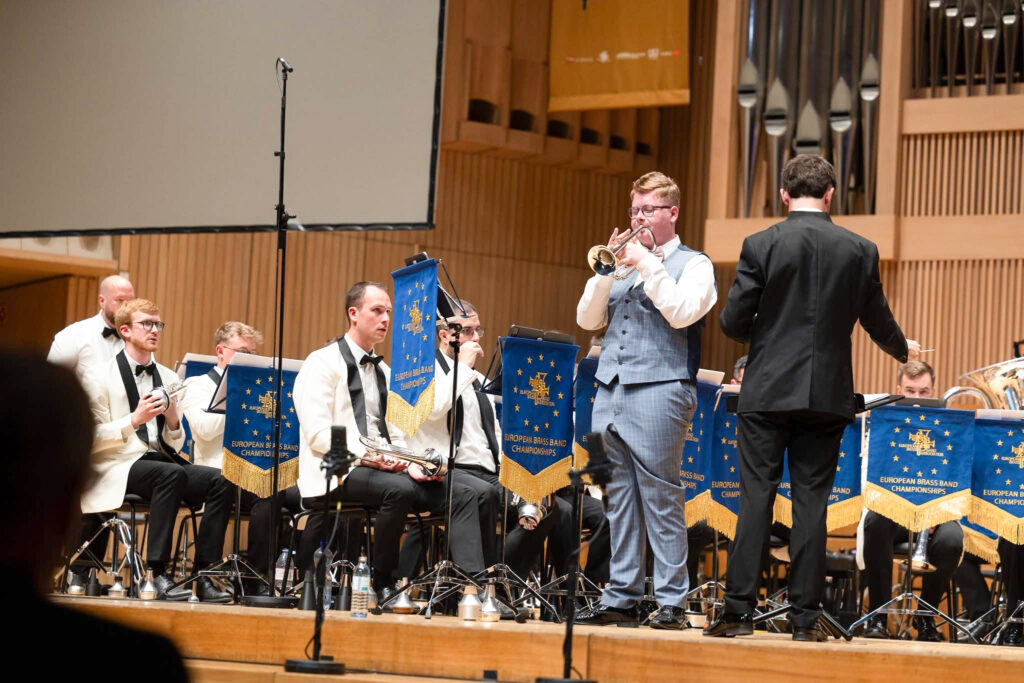 Simon Gabriel se produit sur scène avec son cornet, accompagné d'un brass band (celui-ci et le chef sont visibles en arrière-plan).