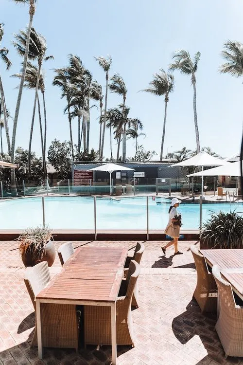 Palm trees and umbrellas line a luxury pool and a woman walks past carrying a bag.