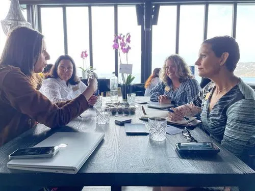 Four people having a conversation, around a table with paper and water. Large windows in the background