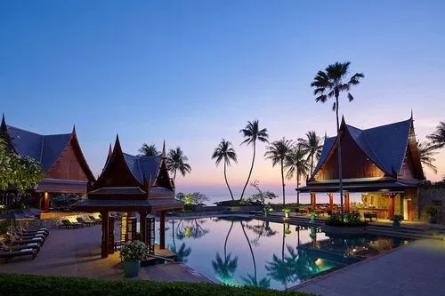 Tropical resort poolside at dusk with traditional architectural structures and palm trees.