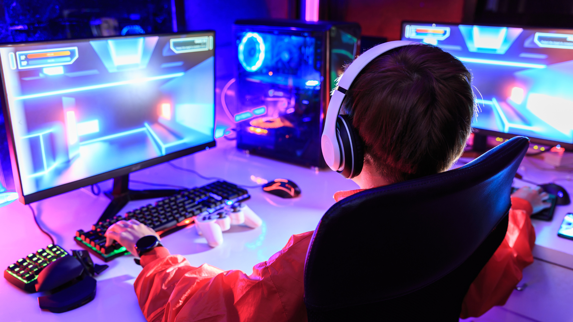 Person with short dark hair wearing white headphones and a red hoodie is sat at a desk gaming on his computer. The photo has bright, neon pink, blue and purple hues.