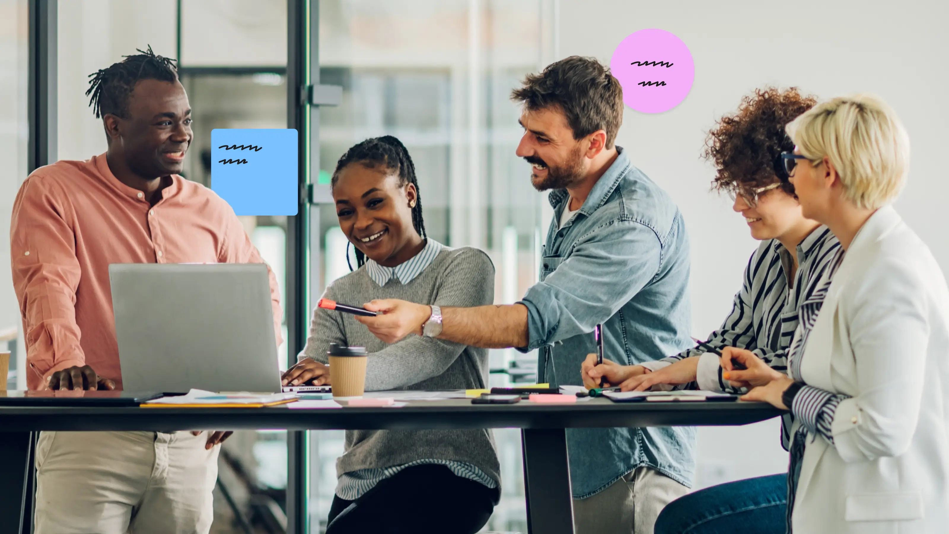 Five colleagues smiling and collaborating around a table with a laptop and documents in an office.