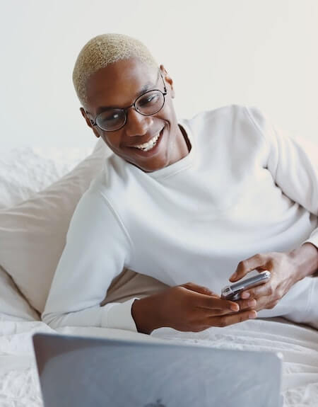 A person looking at a Macbook while holding a phone while smiling.