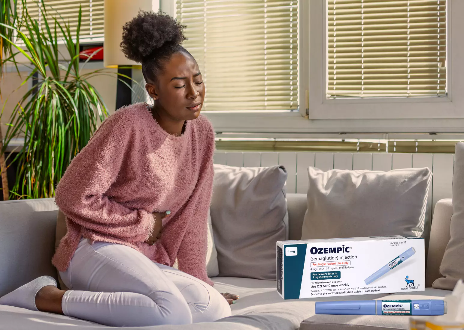 Someone with black hair tied in a bun, sitting on a couch in a pink sweater, holding their stomach. Box of ozempic overlaid