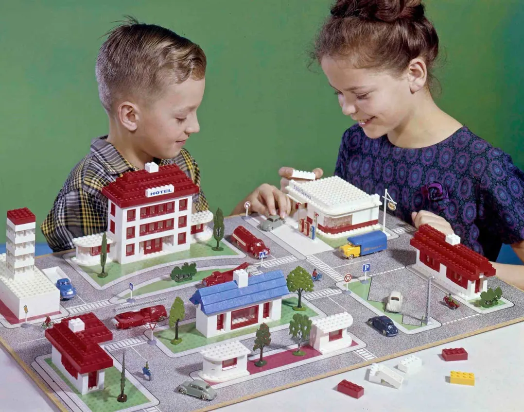 A historical photo of two children, a boy and a girl, playing with an extensive LEGO town layout including multiple buildings, vehicles, and street settings, depicting an early LEGO play experience.