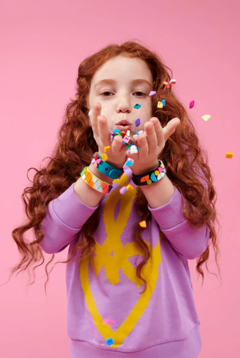 A young girl with curly red hair, wearing purple and yellow clothes, blowing colorful LEGO DOTS confetti from her hands towards the camera, with wristbands adorning both wrists.