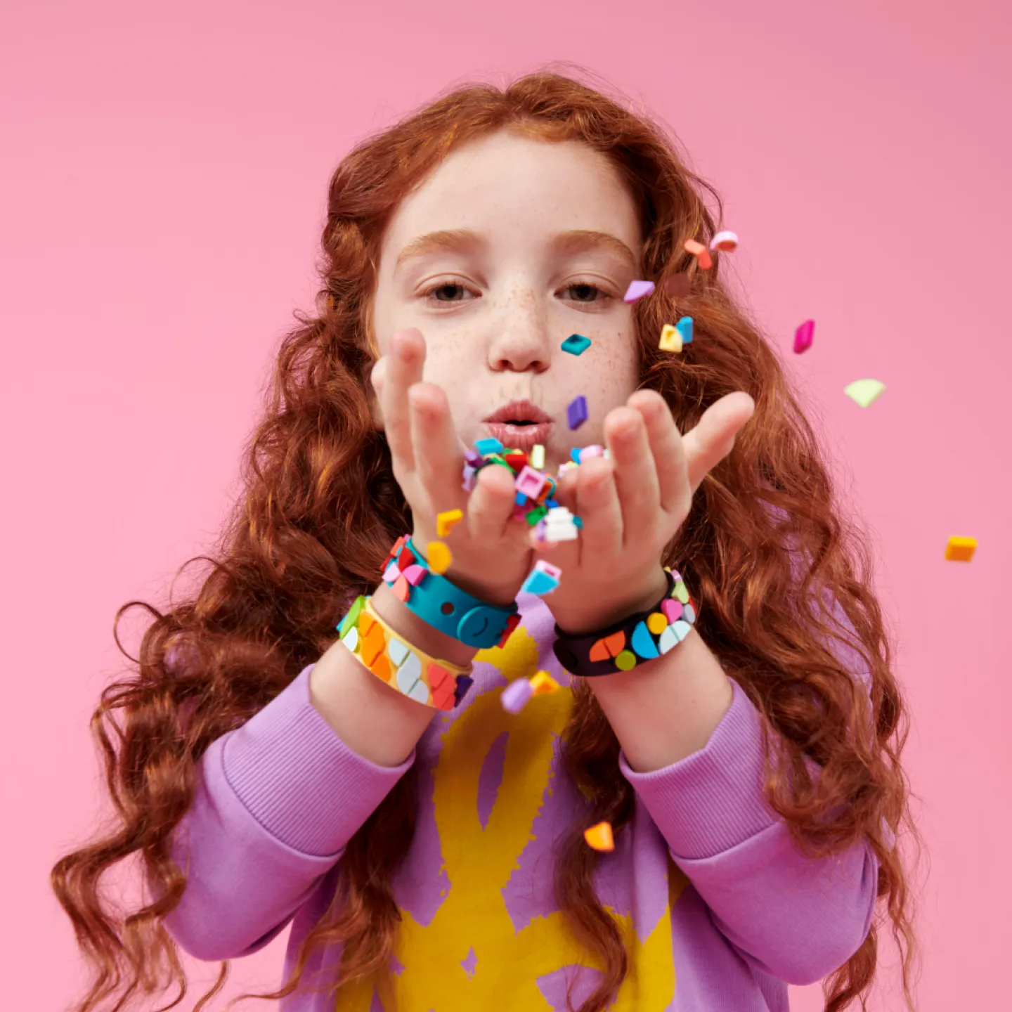 A young girl with curly red hair, wearing purple and yellow clothes, blowing colorful LEGO DOTS confetti from her hands towards the camera, with wristbands adorning both wrists.