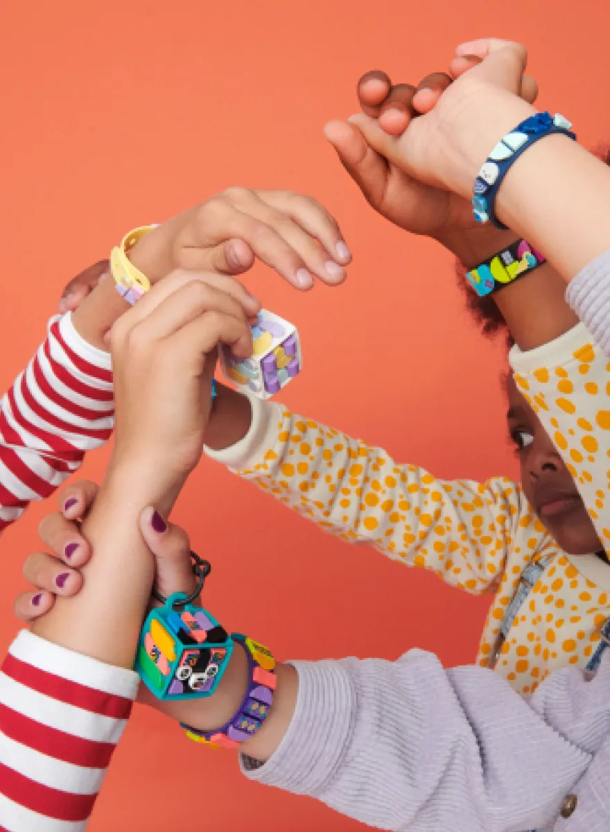 Four children's hands connect in the center of the image against a coral background. Each hand wears a unique LEGO DOTS bracelet with various colorful patterns. One bracelet has pastel colors, another has blue with emoticon tiles, the third features a black base with vibrant graphics, and the fourth has yellow with dot patterns. The image captures a sense of fun and creativity shared among friends.
