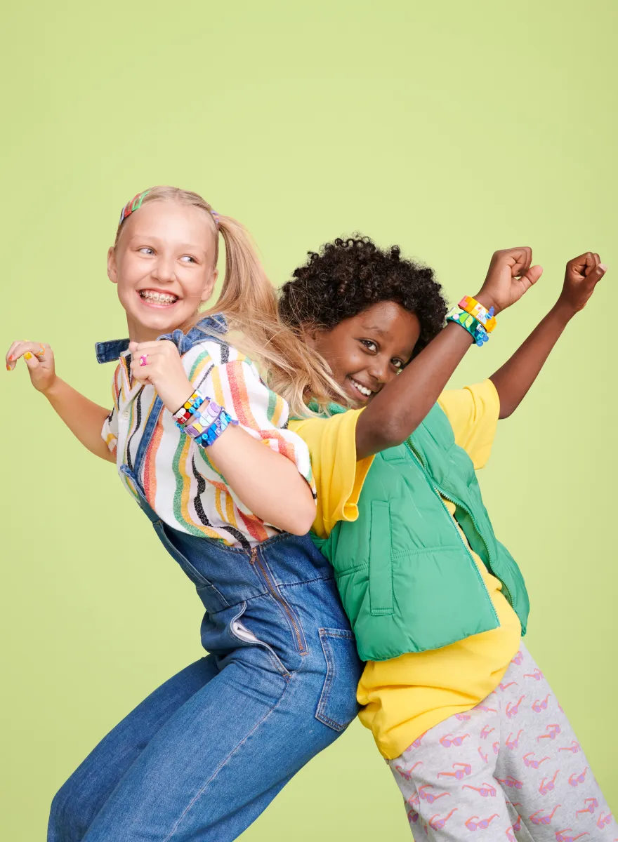 Two children back-to-back, one with blonde hair and the other with curly black hair, smiling and raising their arms in victory, each wearing multiple LEGO DOTS wristbands.