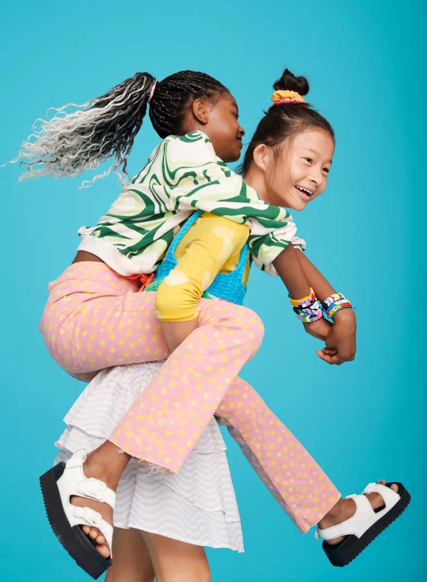 A playful scene with two young girls mid-movement, one carrying the other on her back. The girl being carried has long, flowing, braided hair and a joyous expression, while the carrier, smiling widely, looks over her shoulder. They both wear vibrant, colorful clothing and LEGO DOTS bracelets, showcasing a fun and active moment between friends.