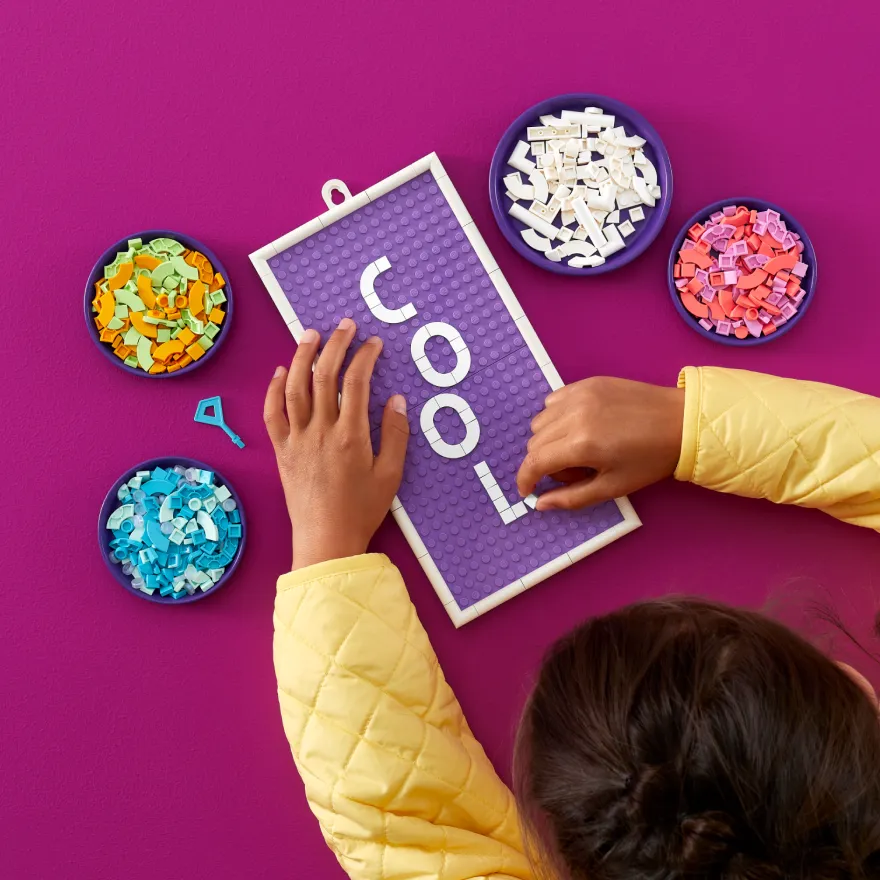 A child's hands placing white letter tiles spelling "COOL" on a purple LEGO DOTS board, with various colored tiles sorted into bowls around the board.