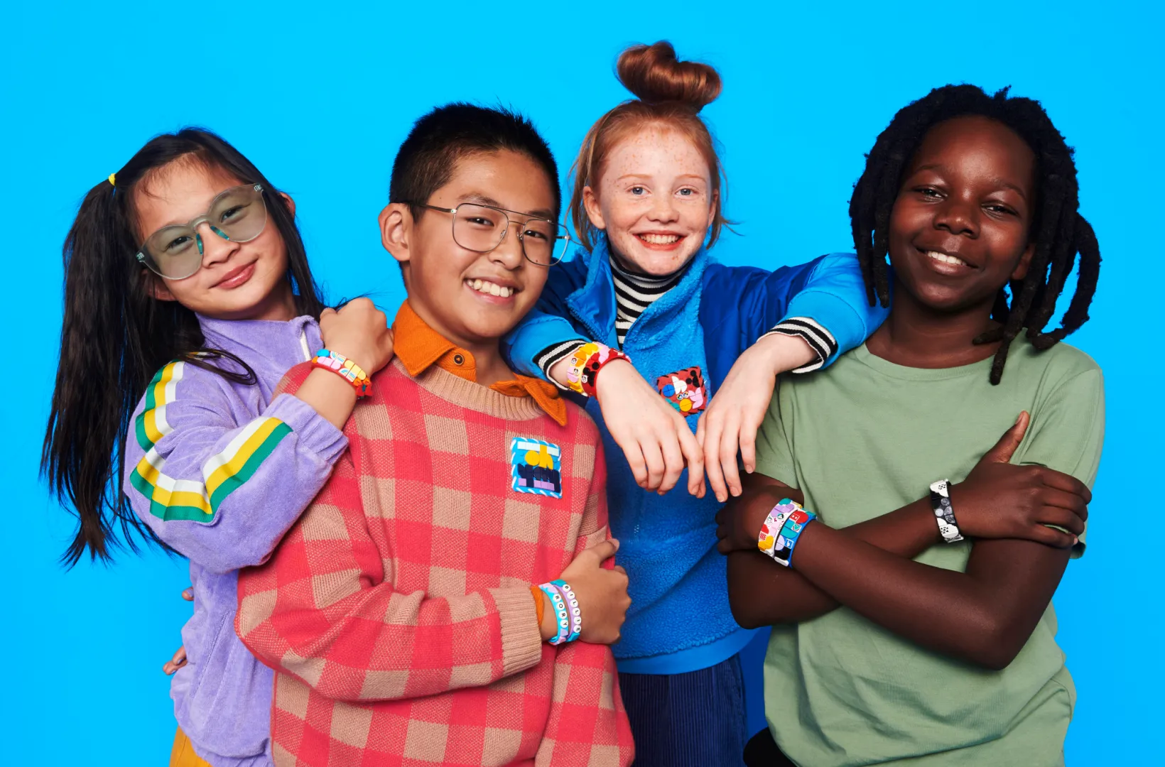 Three children with joyful expressions, standing against a bright blue background. They are each wearing colorful outfits and LEGO DOTS bracelets. The girl on the left has long hair and glasses, the boy in the center has short hair and glasses, and the girl on the right has braided hair.