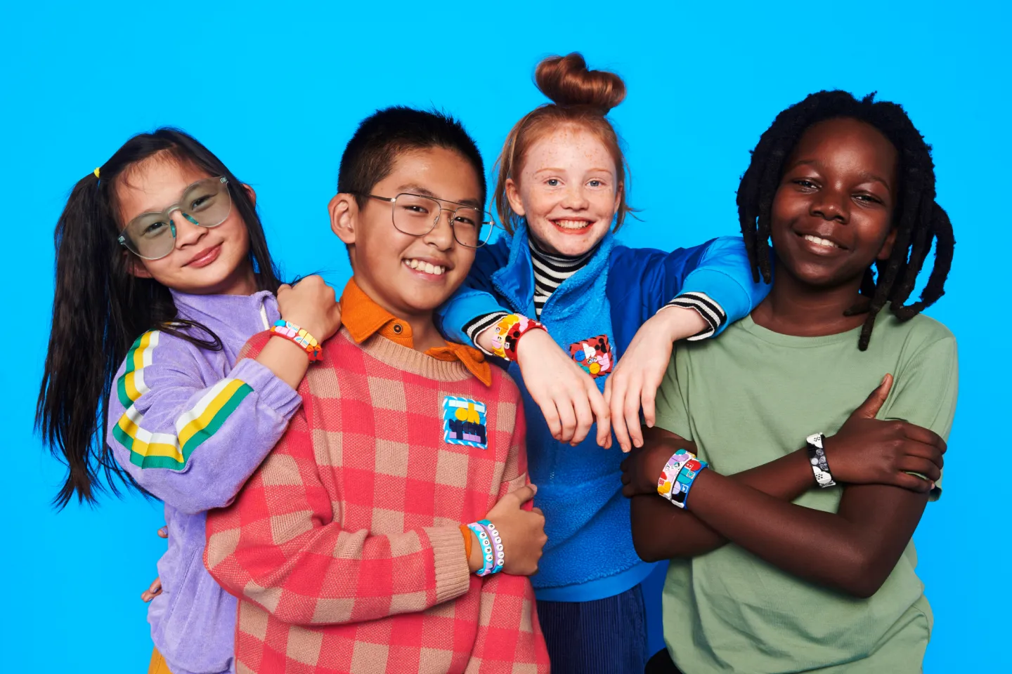 Three children with joyful expressions, standing against a bright blue background. They are each wearing colorful outfits and LEGO DOTS bracelets. The girl on the left has long hair and glasses, the boy in the center has short hair and glasses, and the girl on the right has braided hair.