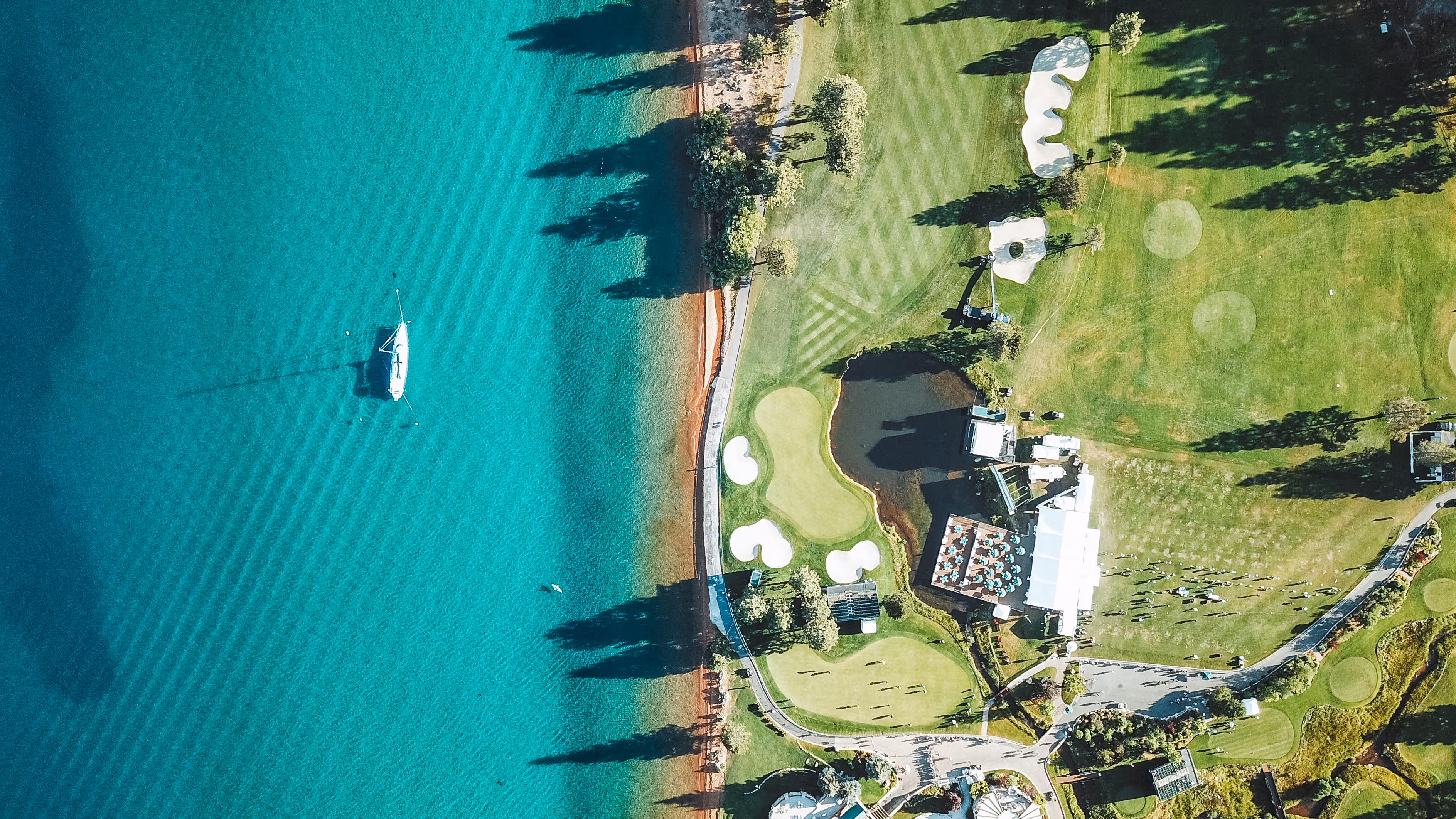 Aerial view of a golf course next to turquoise water with a white sailboat near the shore.