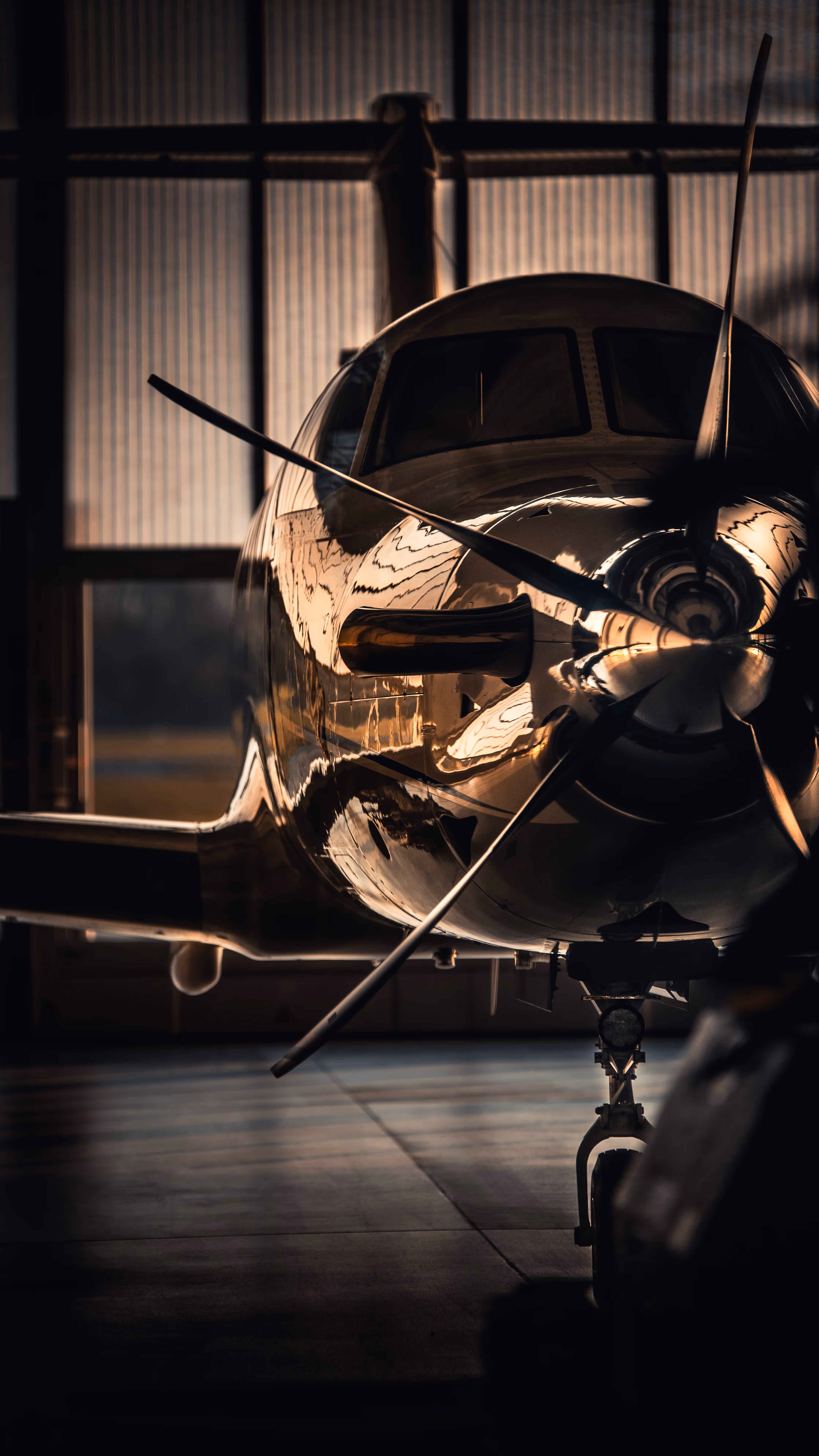 Front view of a propeller airplane inside a dimly lit hangar with sunlight reflections on its polished surface.
