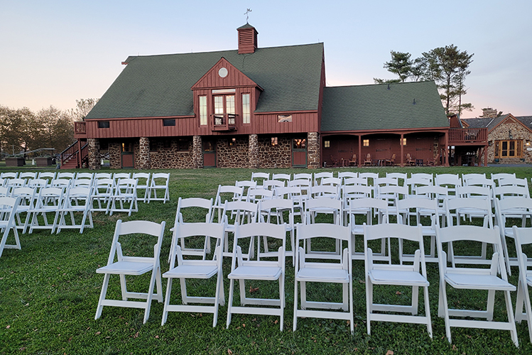 Rows of white folding chairs set on grass in front of a large rustic barn with stone and wood exterior under a clear sky.