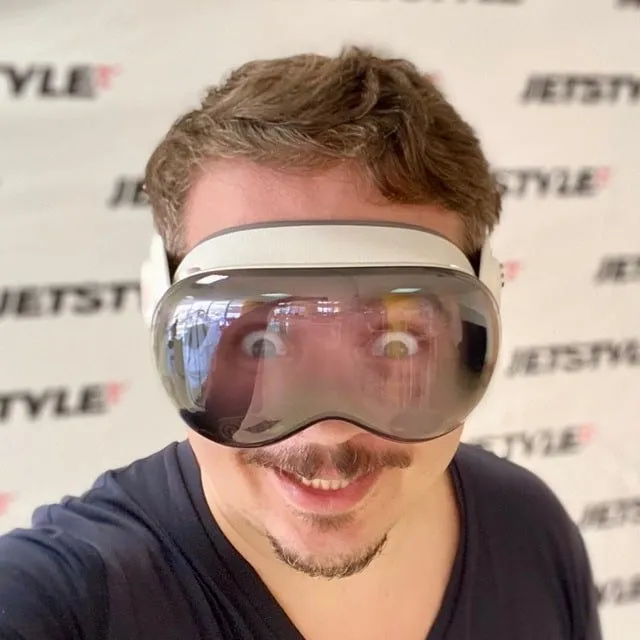 Man with short brown hair and goatee wearing a large virtual reality headset and smiling with wide eyes.