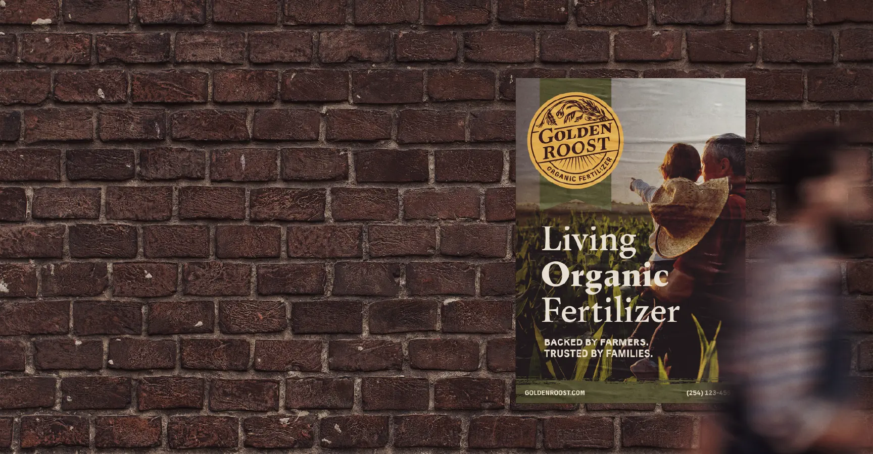 Poster on brick wall showing an adult carrying a child wearing a straw hat in a field with text promoting Golden Roost organic fertilizer.