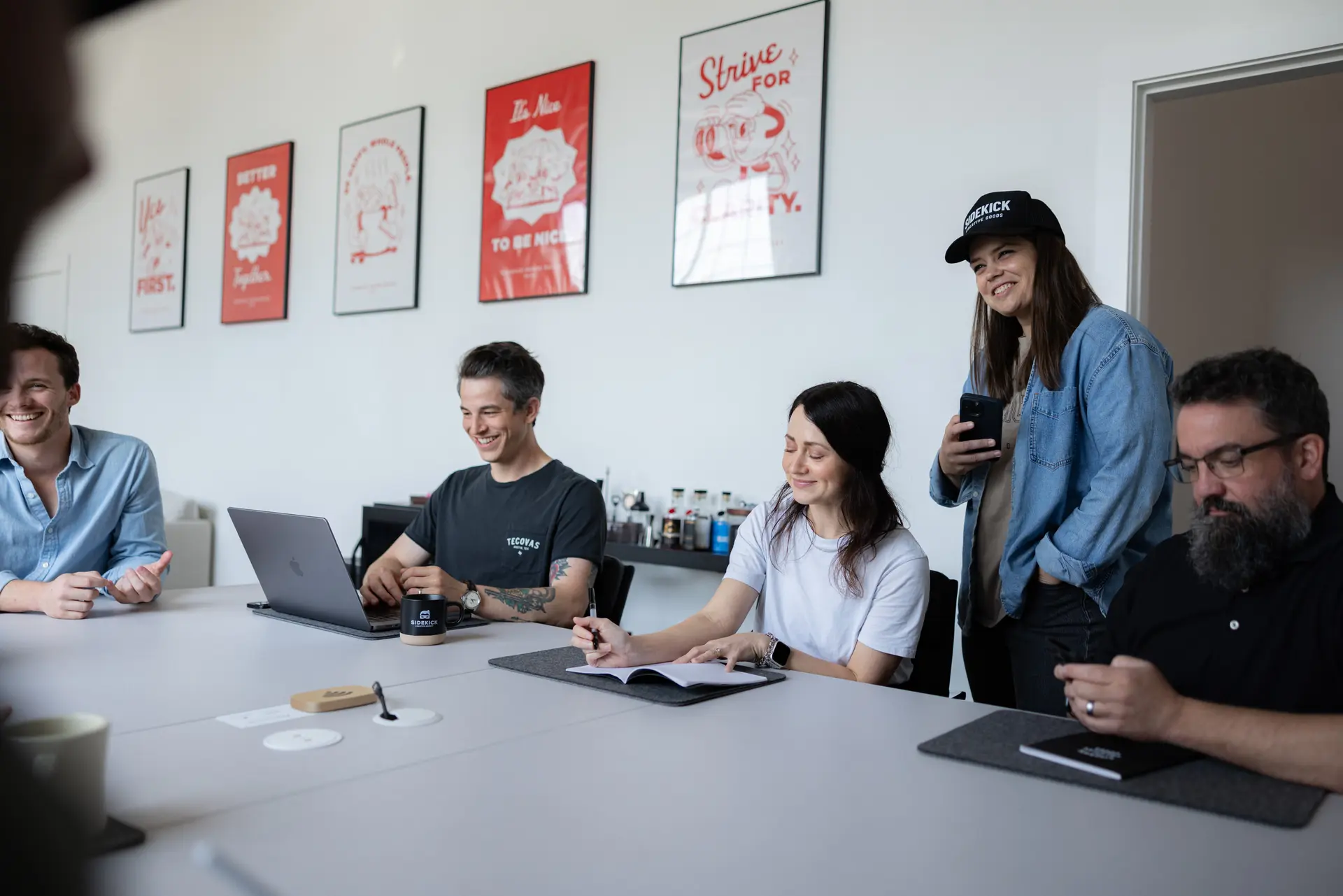 Five people sitting and standing around a conference table in a bright office, smiling and engaged in discussion.