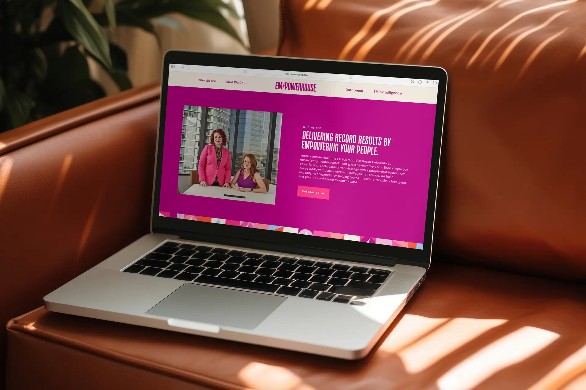 Laptop displaying EMPOWERHOUSE website with two women in a photo and text about empowering people, placed on a brown leather couch.