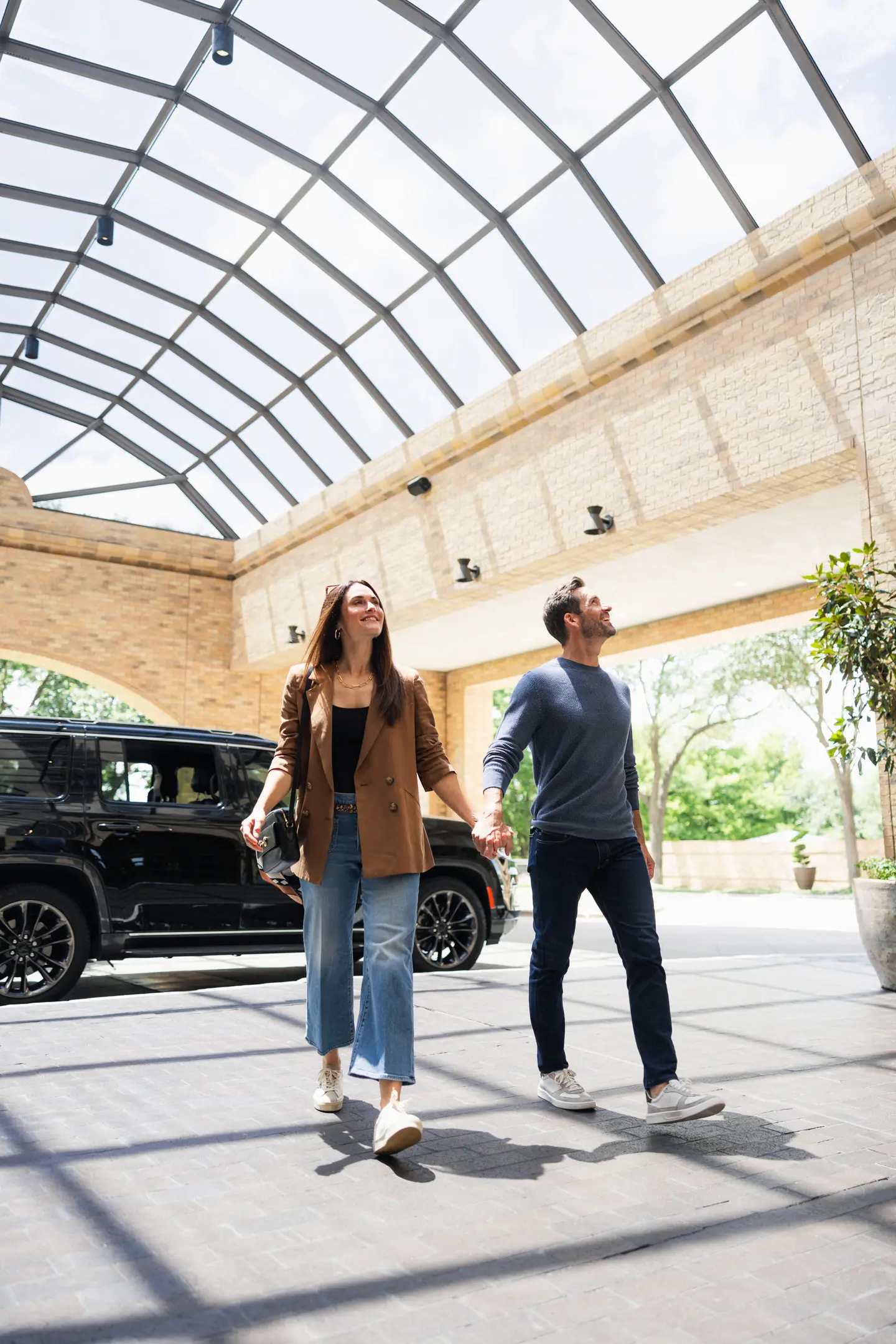 Couple holding hands walking under a glass ceiling with a black SUV parked nearby.