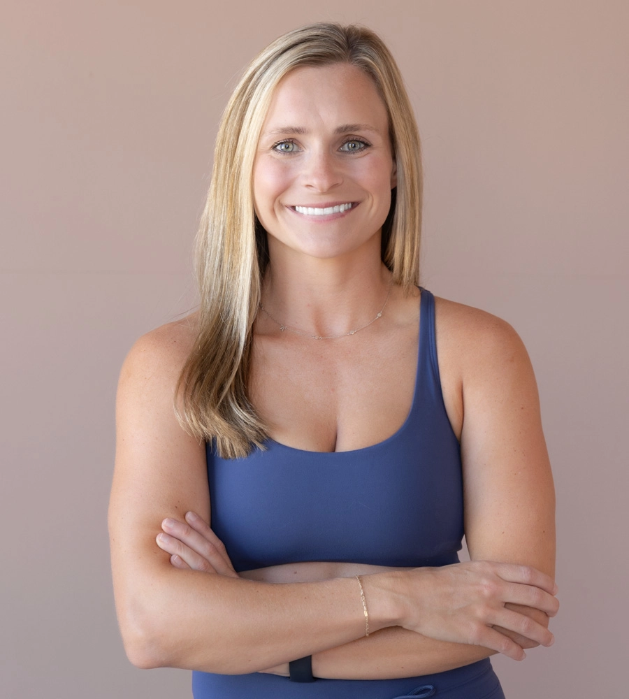 Smiling woman with long blonde hair wearing a blue sleeveless top, arms crossed against a beige background.