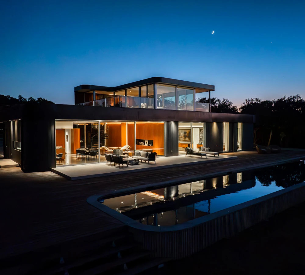 Modern two-story house illuminated at dusk with large glass windows and a reflecting pool in the foreground.